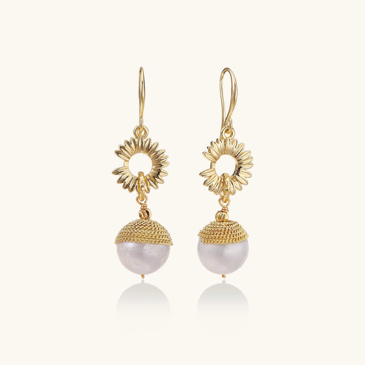 Gold sunburst charm earrings with baroque pearl drops on French hooks against cream background