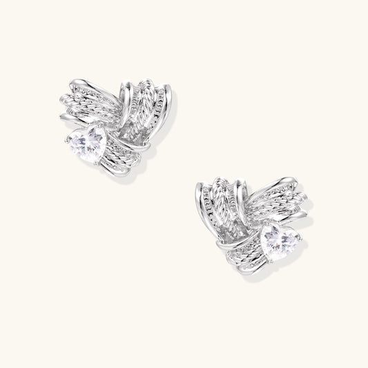 Pair of sculptural white gold-plated stud earrings with twisted ribbon design and sparkling zirconia stones on cream background